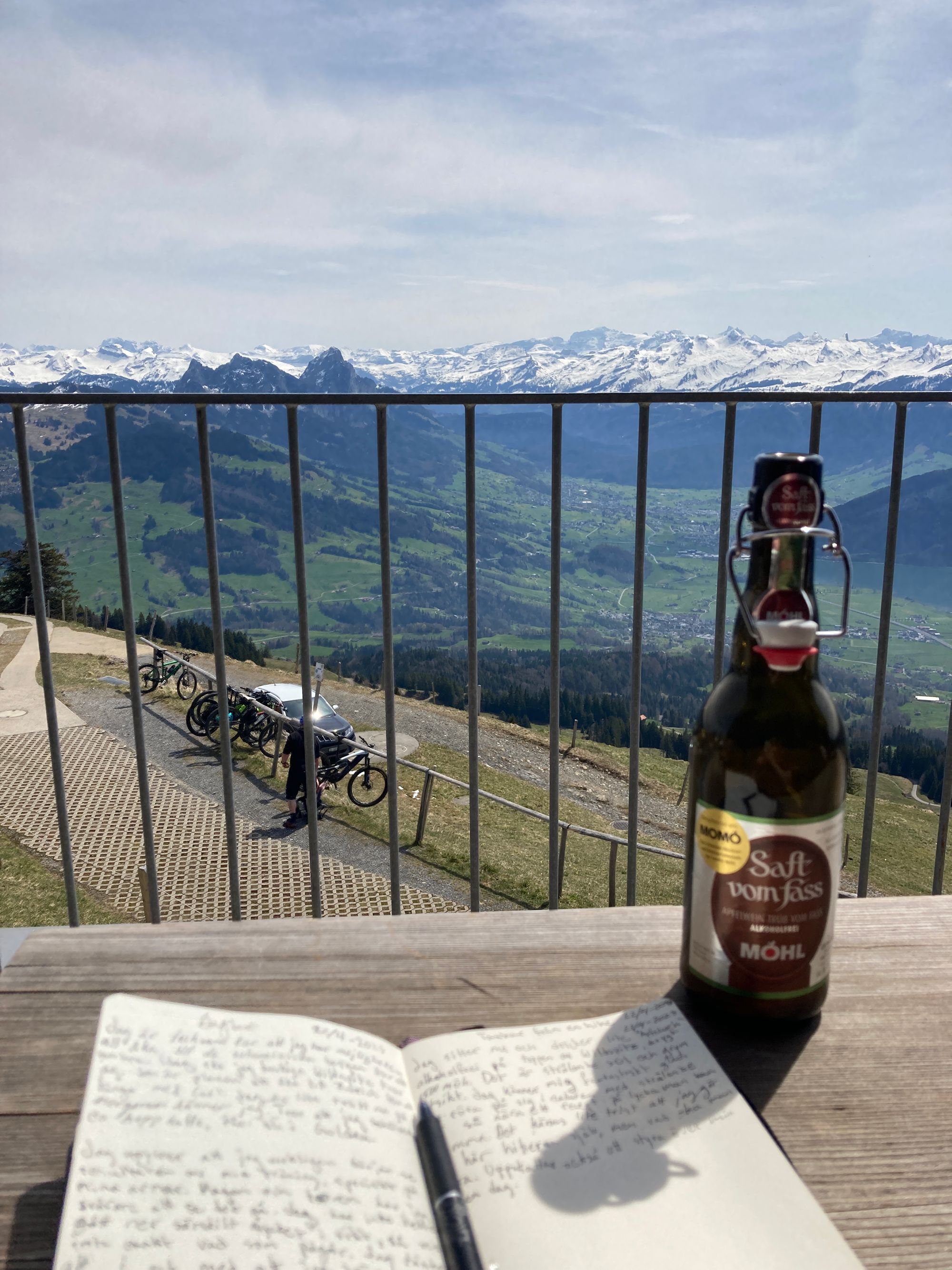 An open journal with a pen, on a table with a bottle next to it. The background is an alpine valley and snowcapped mountains.