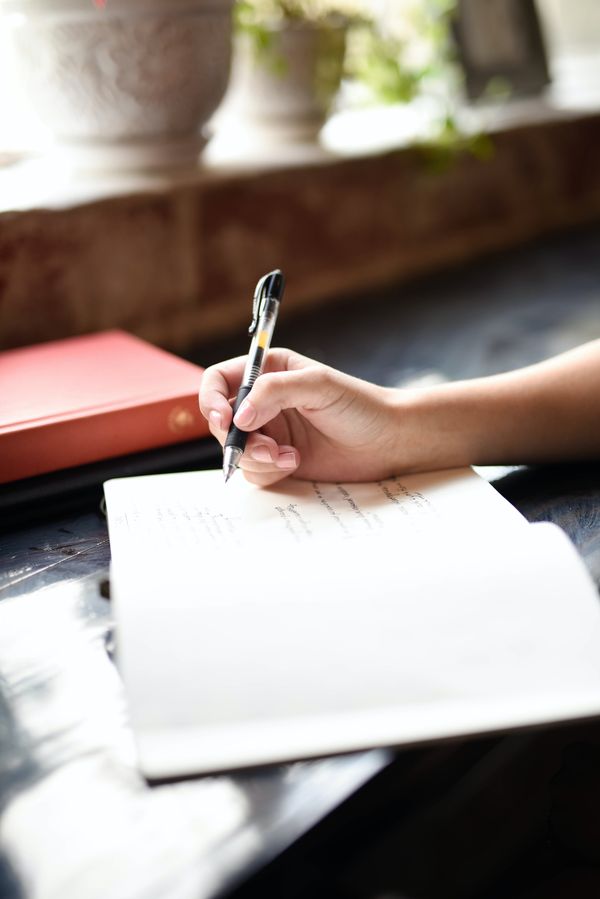 Person writing in a journal at window. You can see the hand with a pen in hand and the open journal.