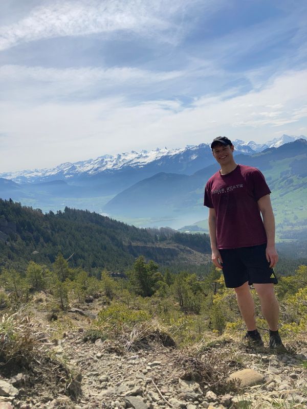 Me standing on a mountain with the alps on the horizon, forest on the slopes and an alpine valley in the background.